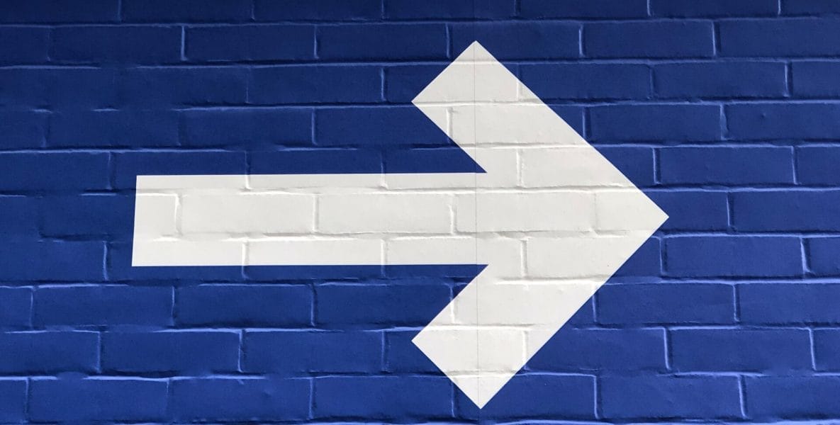 A white arrow on a blue brick wall, directing to the right.